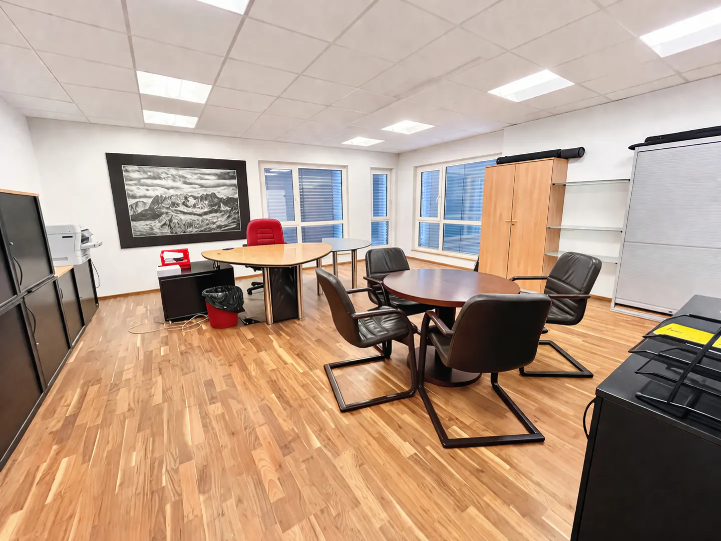 Bright office space with wood floors, a round table with black chairs, and a desk with a red chair. A mountain picture hangs on the wall.