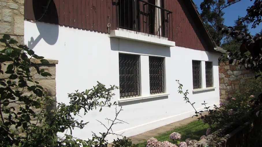 A two-story house with white walls, a brown roof, and a small balcony with black railings. Three windows with bars are visible.