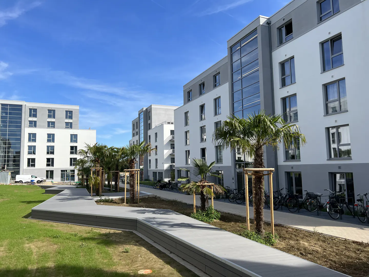 Modern white and gray apartment buildings with palm trees and bike racks under a blue sky.