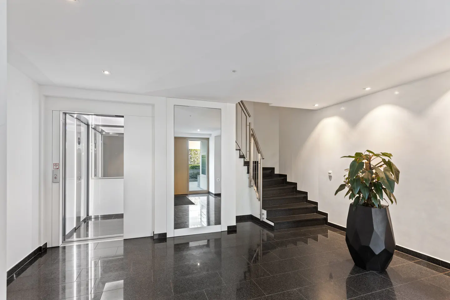 Bright lobby with black marble floors, white walls, and recessed lighting. An elevator, stairs, and a potted plant are visible.