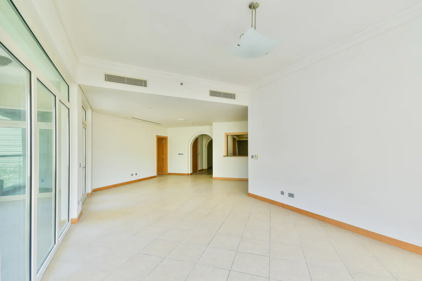 Bright, empty room with white walls, tile floor, and large windows. A doorway leads to another room. A light fixture hangs from the ceiling.