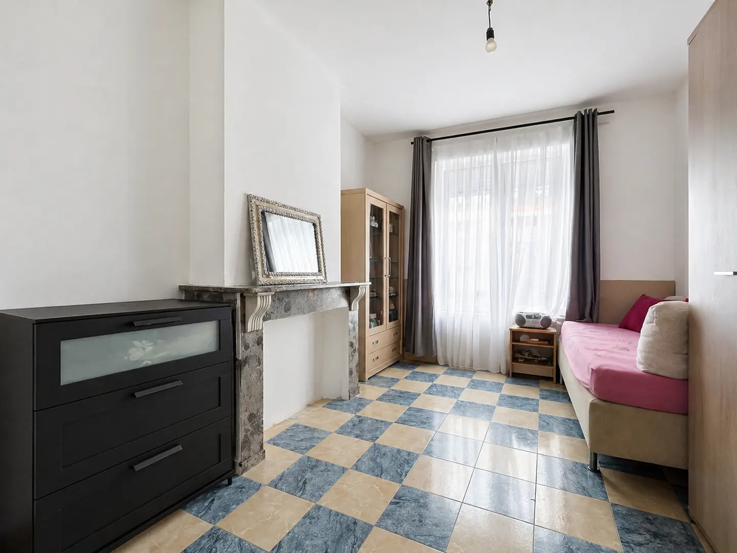 A bright room with blue and beige checkered tile floor, a black dresser, a marble fireplace, and a daybed with pink sheets.