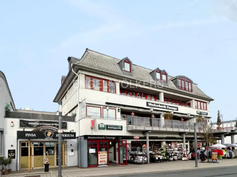 Exterior view of a three-story commercial building with shops and restaurants. The building has a gray roof and white walls. "Engel & Völkers" logo is visible.