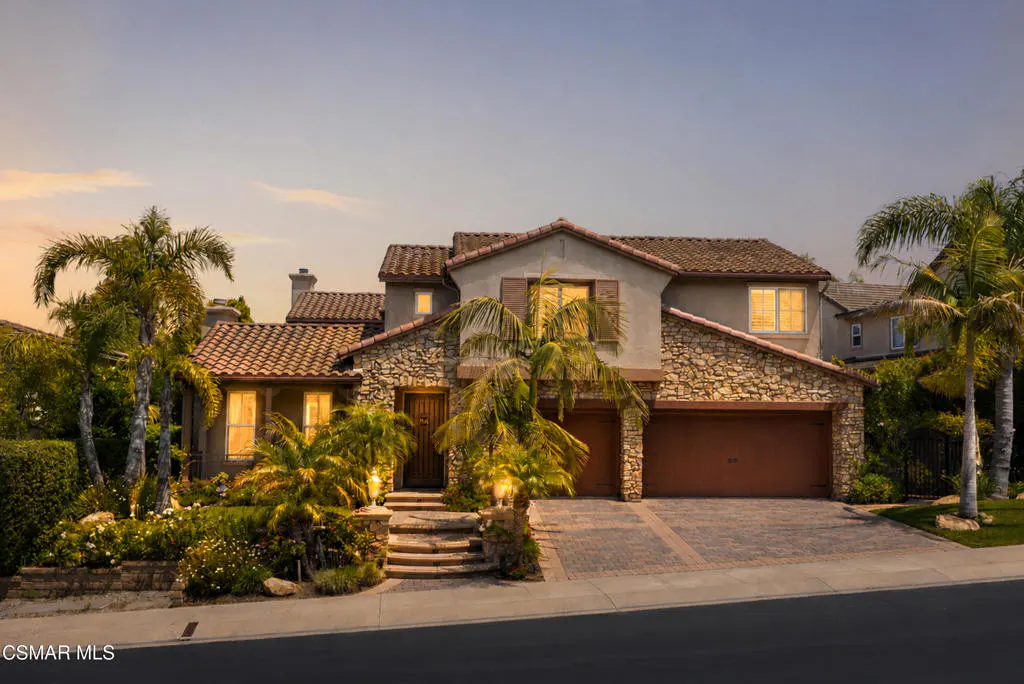 Two-story Mediterranean home with stone facade, tile roof, palm trees, and a paved driveway at sunset.