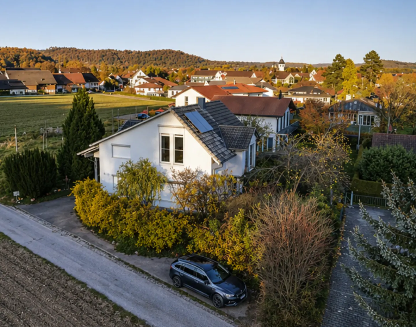 Aerial view of a white house with a gray roof, solar panels, and a gray car parked on the street. Trees and other houses are in the background.