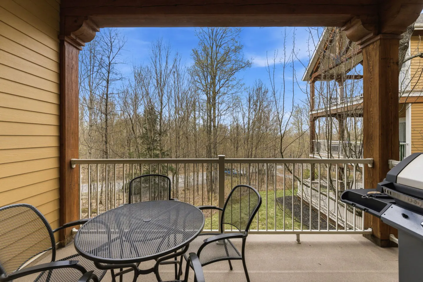 Balcony view with a black metal table and chairs, a grill, and a view of trees and a blue sky.