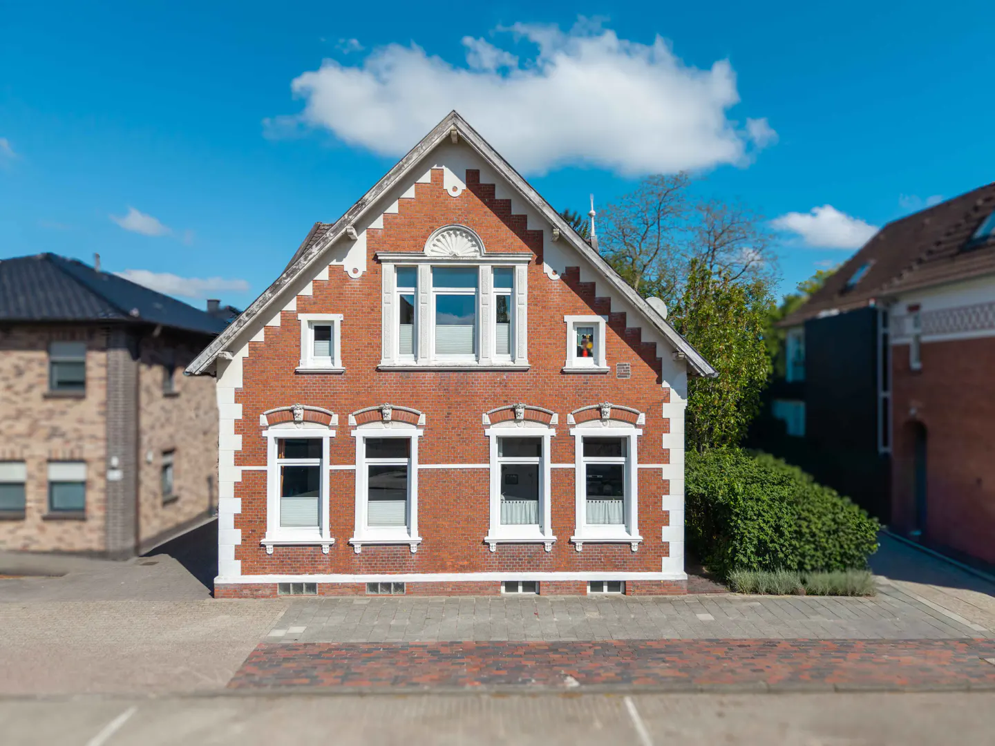 A two-story red brick house with white trim and a gabled roof, situated on a paved street under a bright blue sky.