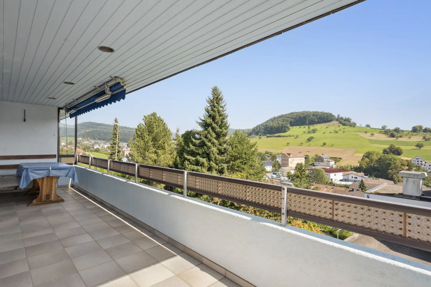 A balcony with a table overlooks a green, hilly landscape under a blue sky.