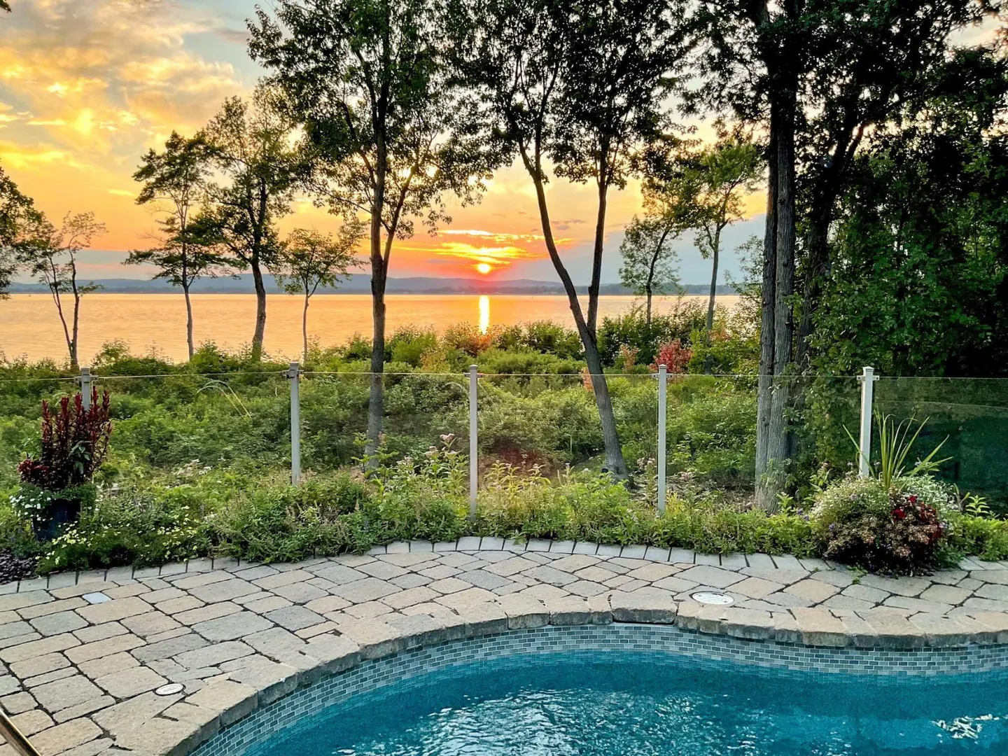 Sunset view from a pool deck. Trees frame the orange sun reflecting on the water. A glass fence separates the pool from the greenery.