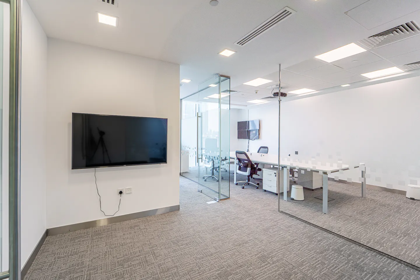 A modern office space with a large TV on the wall, glass-walled meeting room, and white desks.