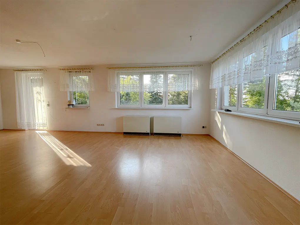 Bright, empty room with wood floors, white walls, and lace curtains on windows. Sunlight streams in.