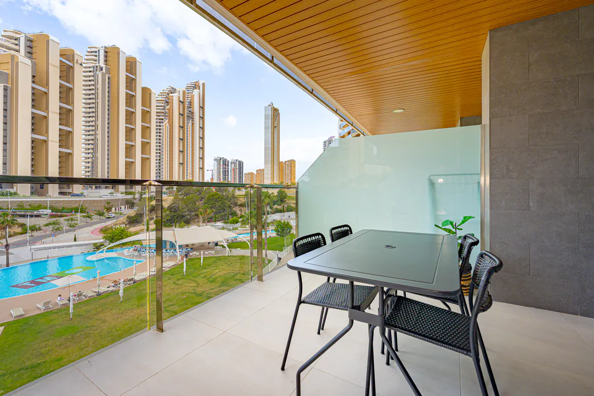 Modern balcony with a dining set overlooking a swimming pool, green lawn, and tall residential towers in the distance.