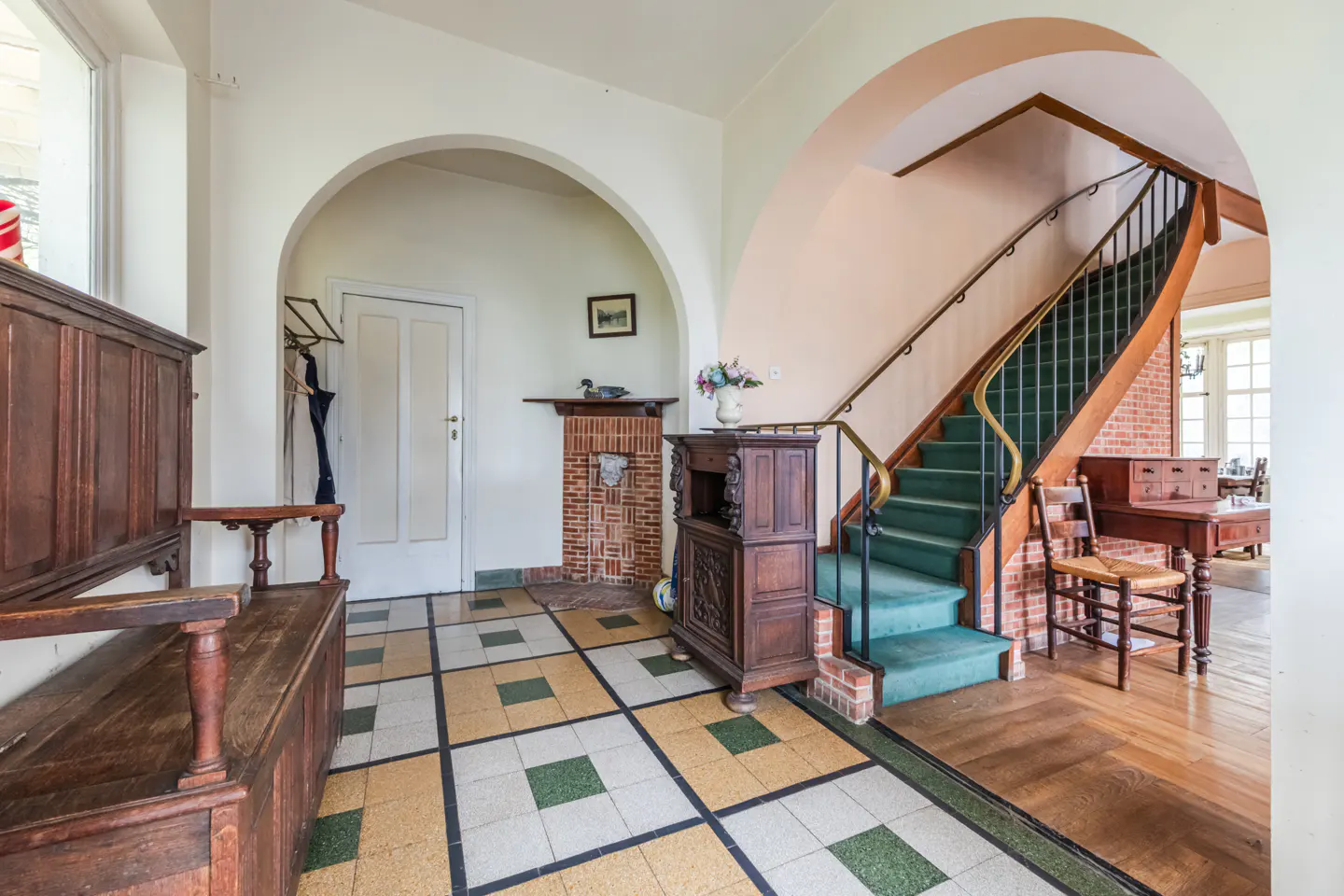 A foyer with a wooden bench, tiled floor, arched doorways, and a staircase with green carpet.