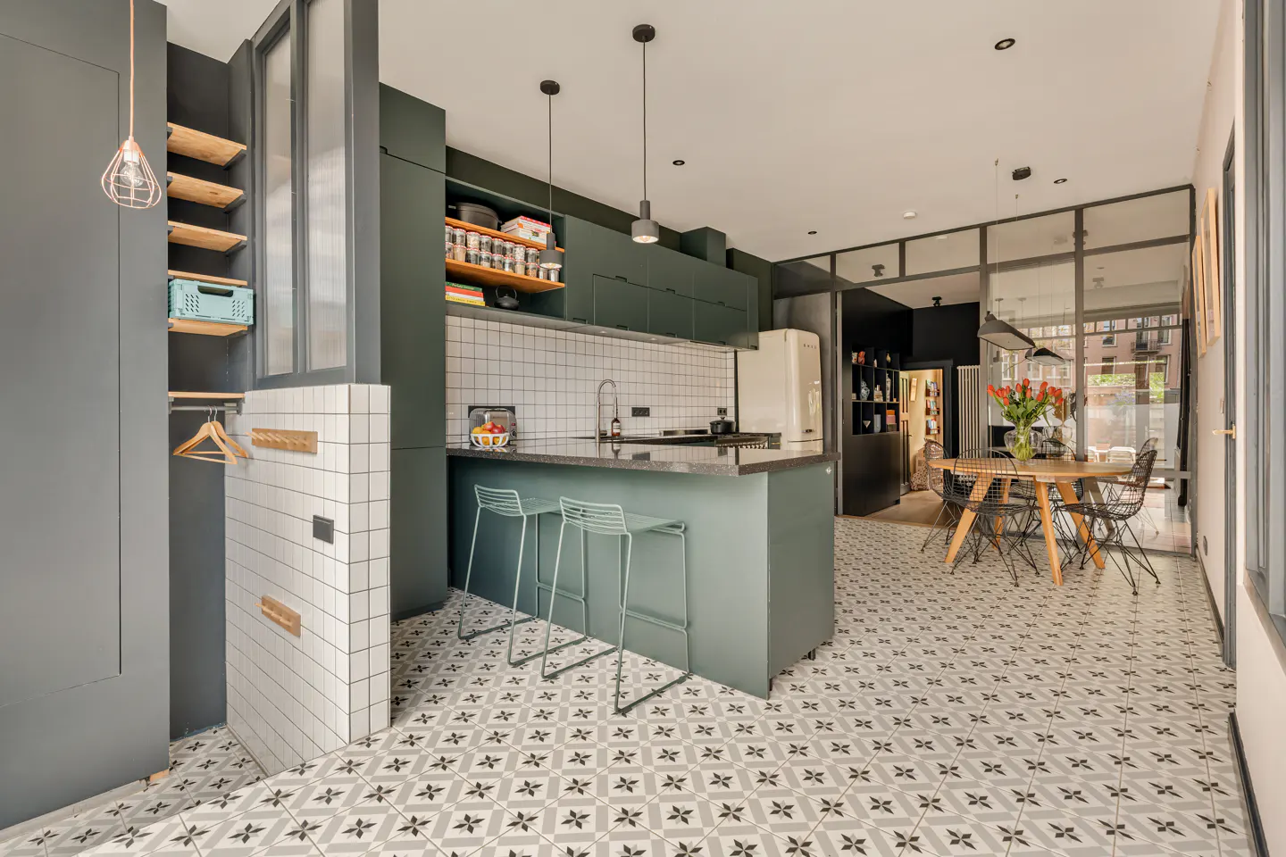 A modern kitchen with dark green cabinets, a white tiled backsplash, and a patterned floor. A dining table with chairs is visible in the background.