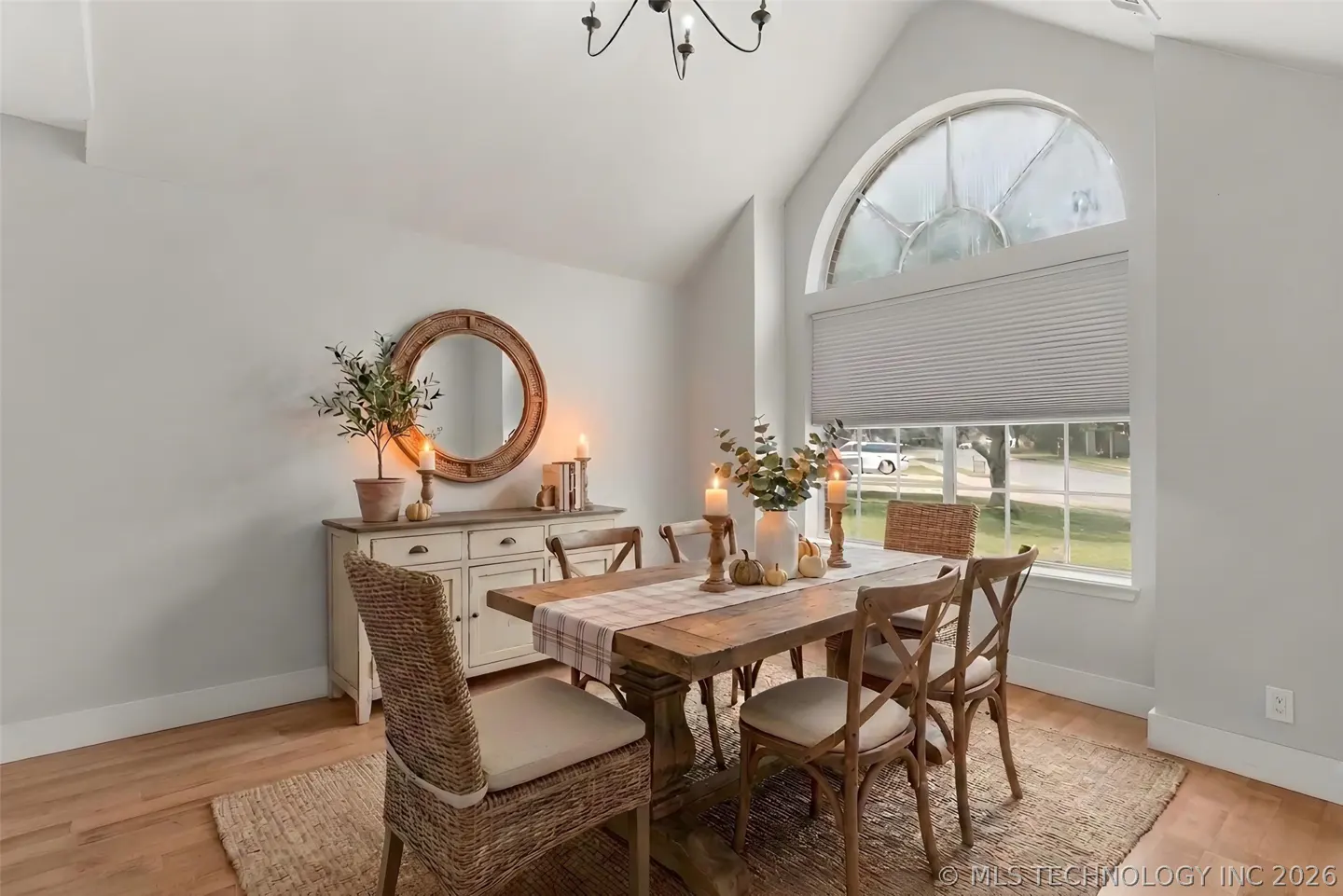 Dining room with wood table, woven chairs, and a plaid runner. Candles and pumpkins decorate the table. A round mirror hangs above a white sideboard.