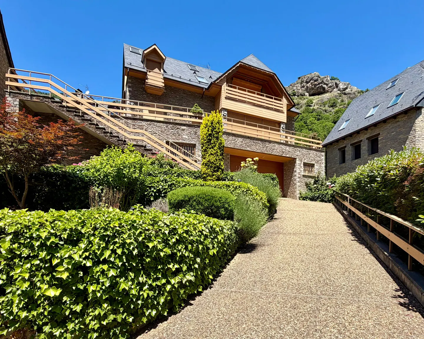 Stone house with wooden balconies and a gravel driveway, surrounded by lush green hedges under a clear blue sky.