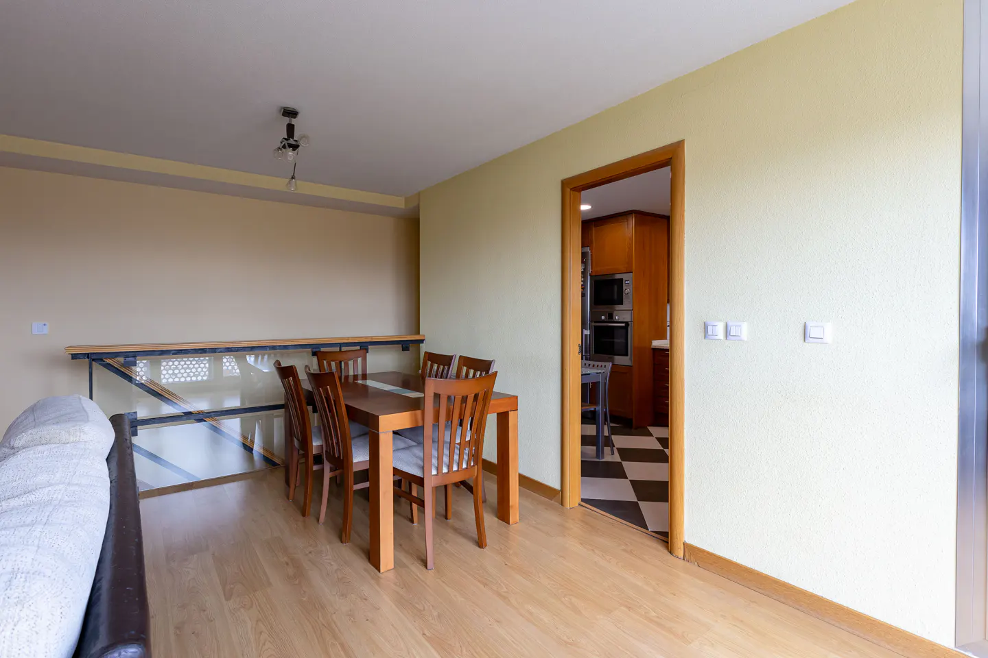 Dining area with wooden table, chairs, and light wood floors. Kitchen visible through doorway with black and white checkered floor.