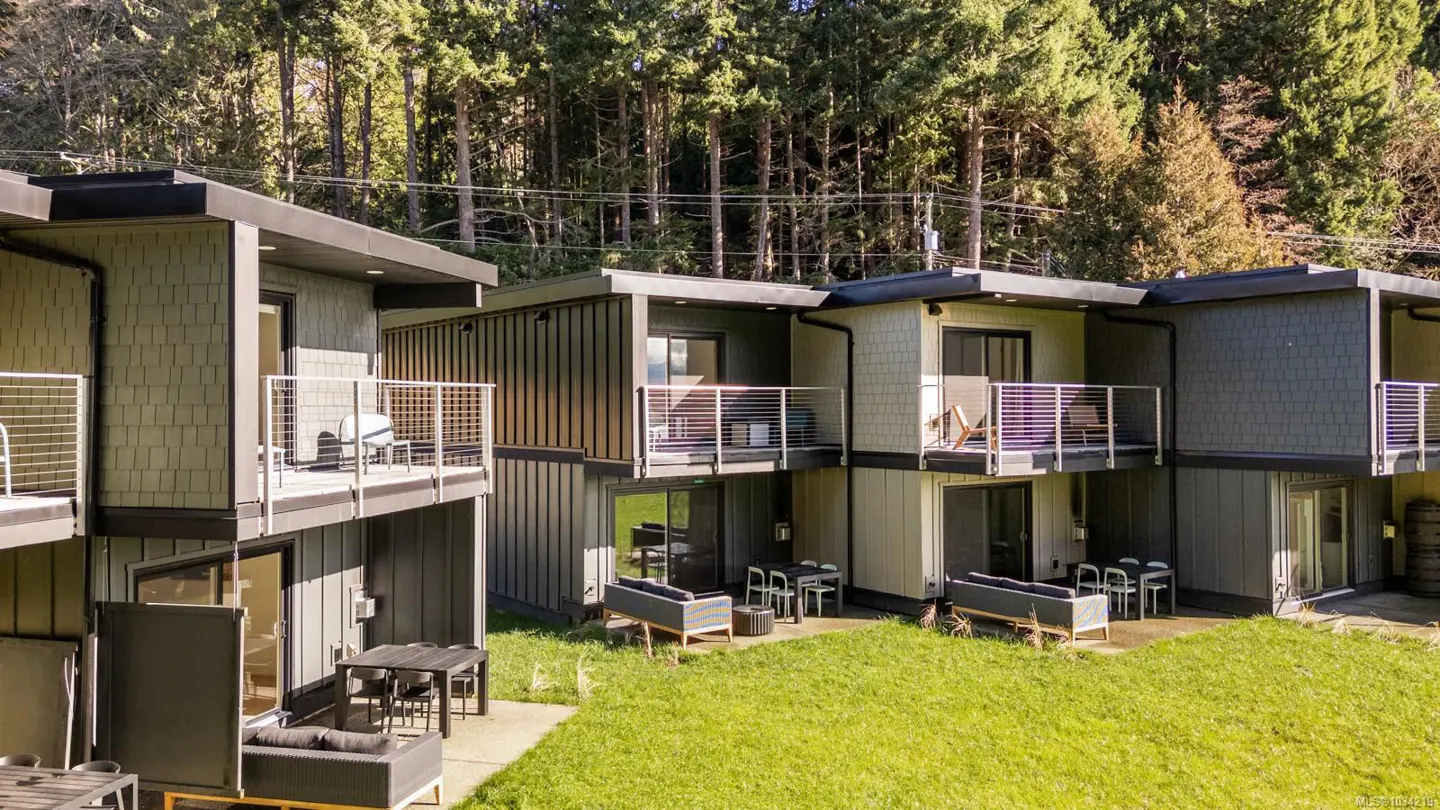 Two-story gray condos with balconies and outdoor seating on a green lawn, backed by a forest.