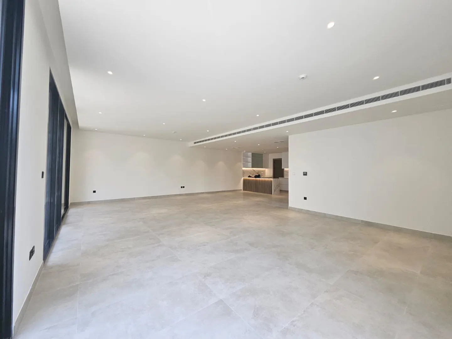 An empty, bright room with gray tile floors and white walls. Large black-framed windows are on the left, and a kitchen is visible in the background.