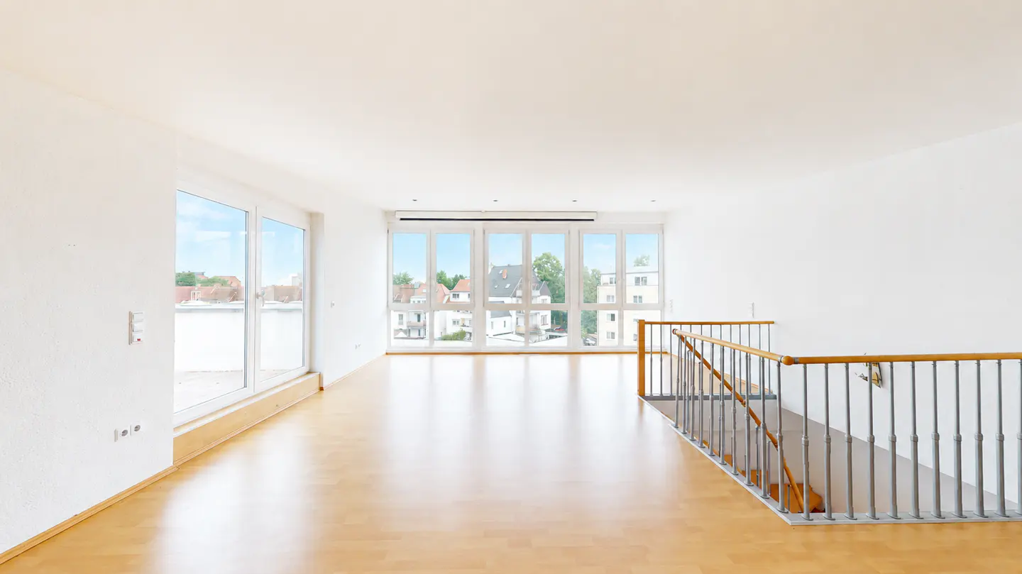 Bright, empty room with wood floors, white walls, and large windows overlooking a cityscape. A staircase with a wooden railing is on the right.