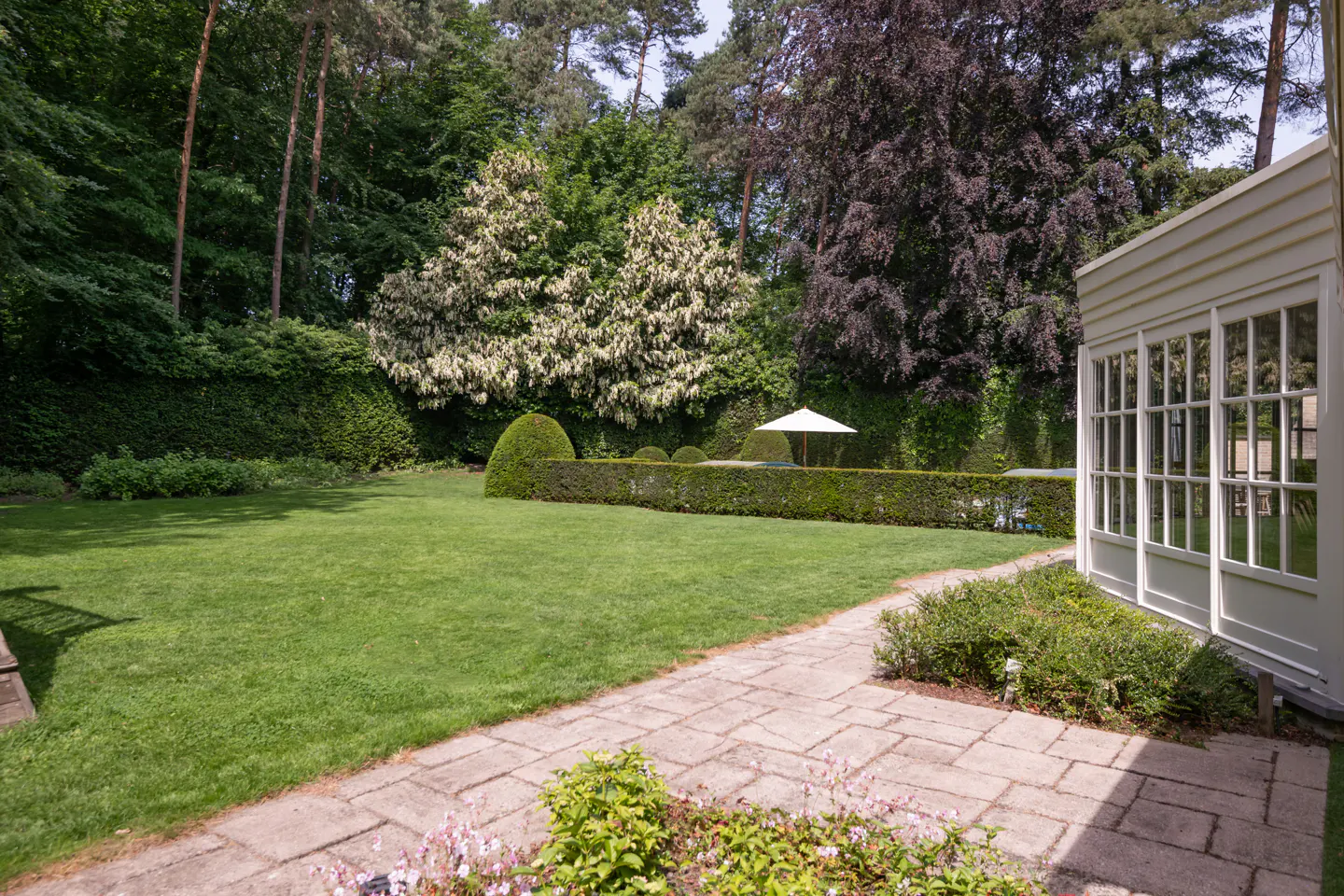 A lush green lawn with manicured hedges and trees, viewed from a stone patio next to a white building.
