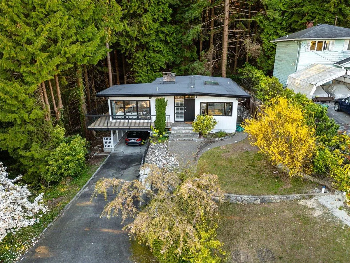 Aerial view of a white house with a black roof, deck, and a car parked underneath, surrounded by lush greenery.