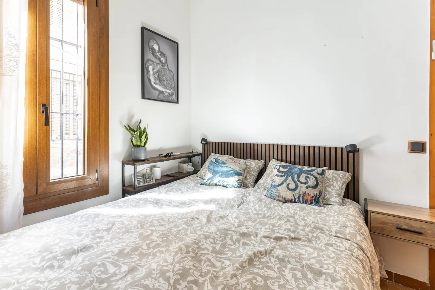 Bedroom with a wooden headboard bed, patterned duvet, and ocean-themed pillows. A window and a black and white picture are on the white walls.