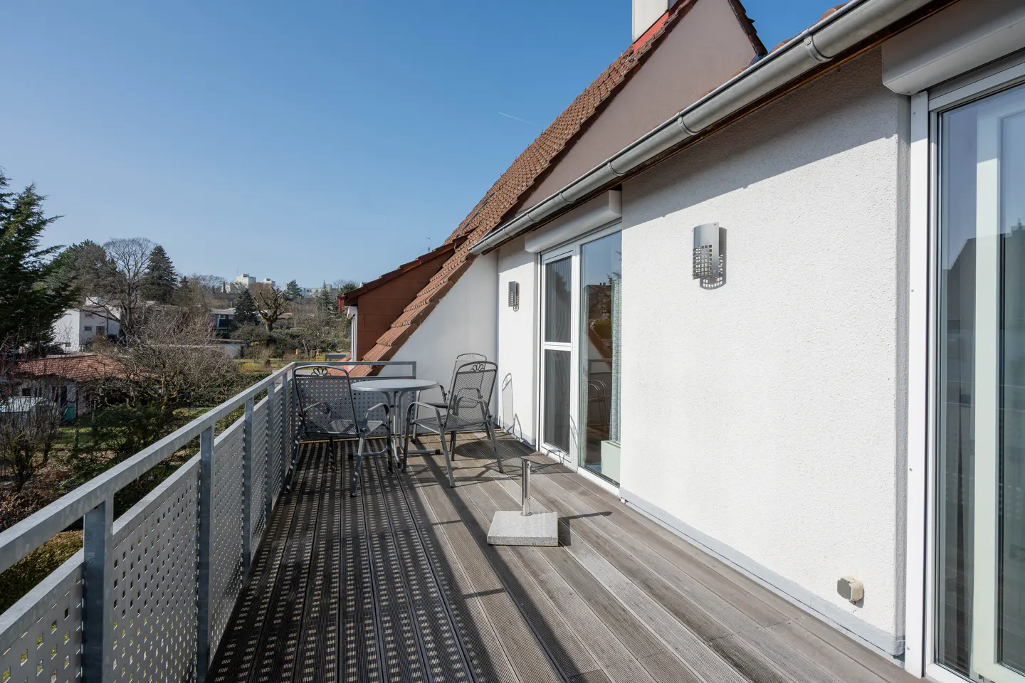 A sunny balcony with a metal table and chairs overlooks a green landscape under a clear blue sky. The balcony has a wooden floor and a metal railing.
