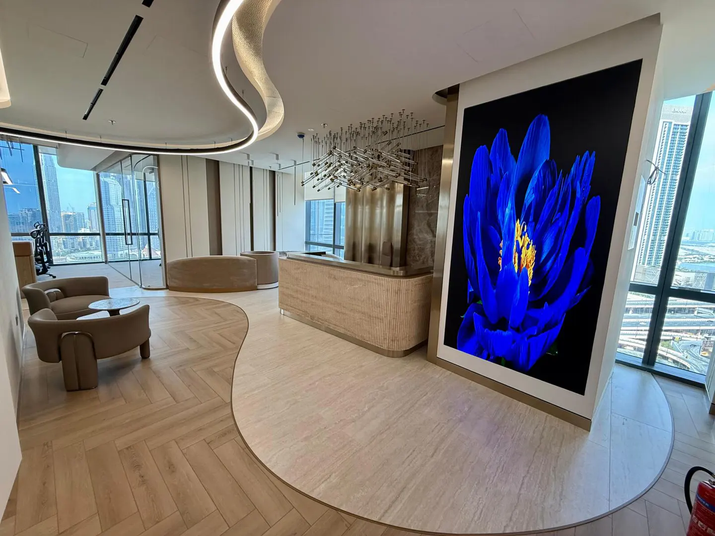 Modern office lobby with beige seating, reception desk, and a large blue flower art piece. Cityscape visible through windows.