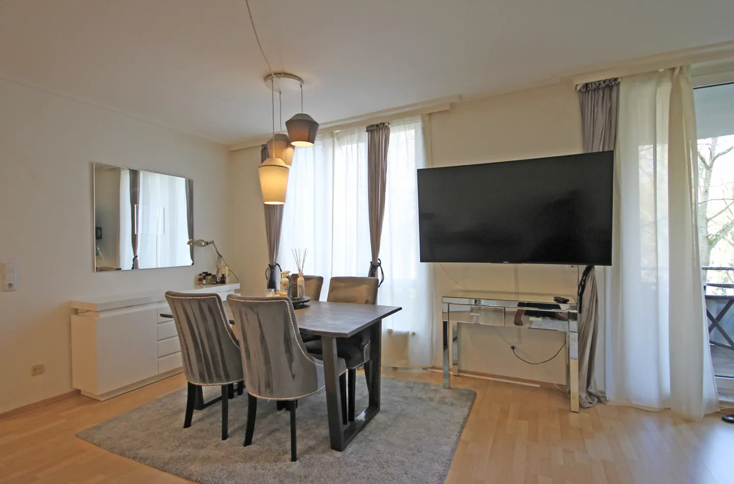 A dining room with a dark wood table, grey velvet chairs, and a large TV on the wall.