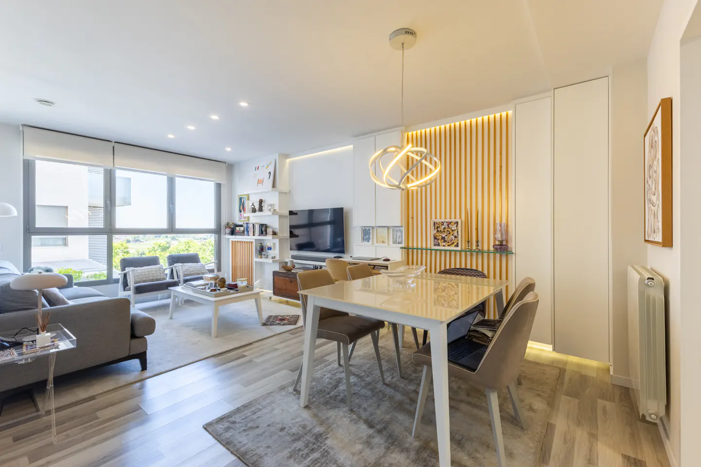 Modern open-plan living and dining area with a white table, grey chairs, and a cozy lounge space with large windows.