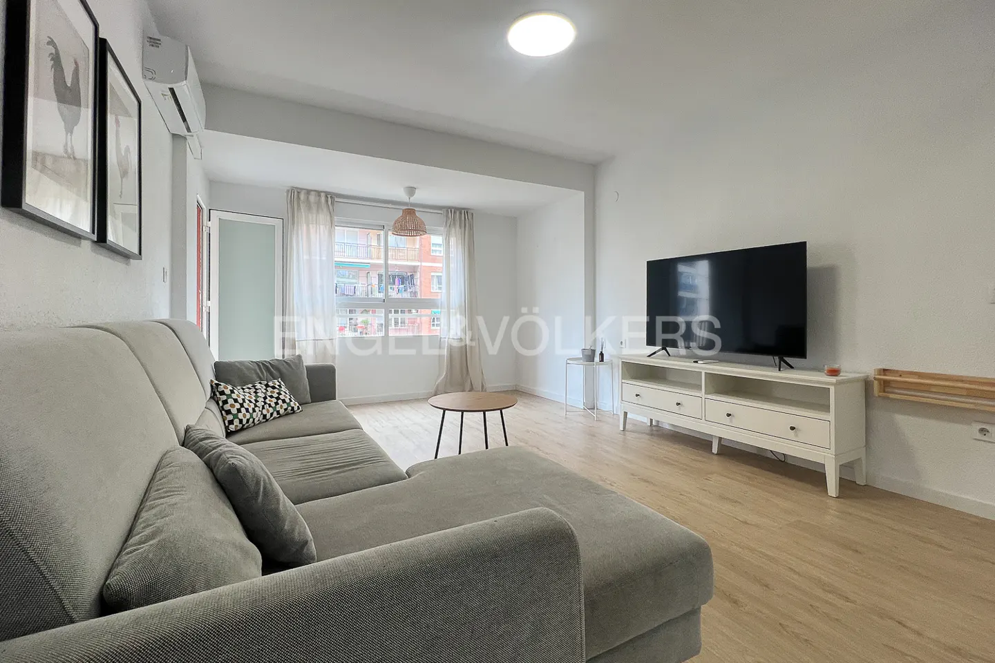 Bright living room with a gray sectional sofa, TV, white console, and wood floors. A window with curtains overlooks a city view.