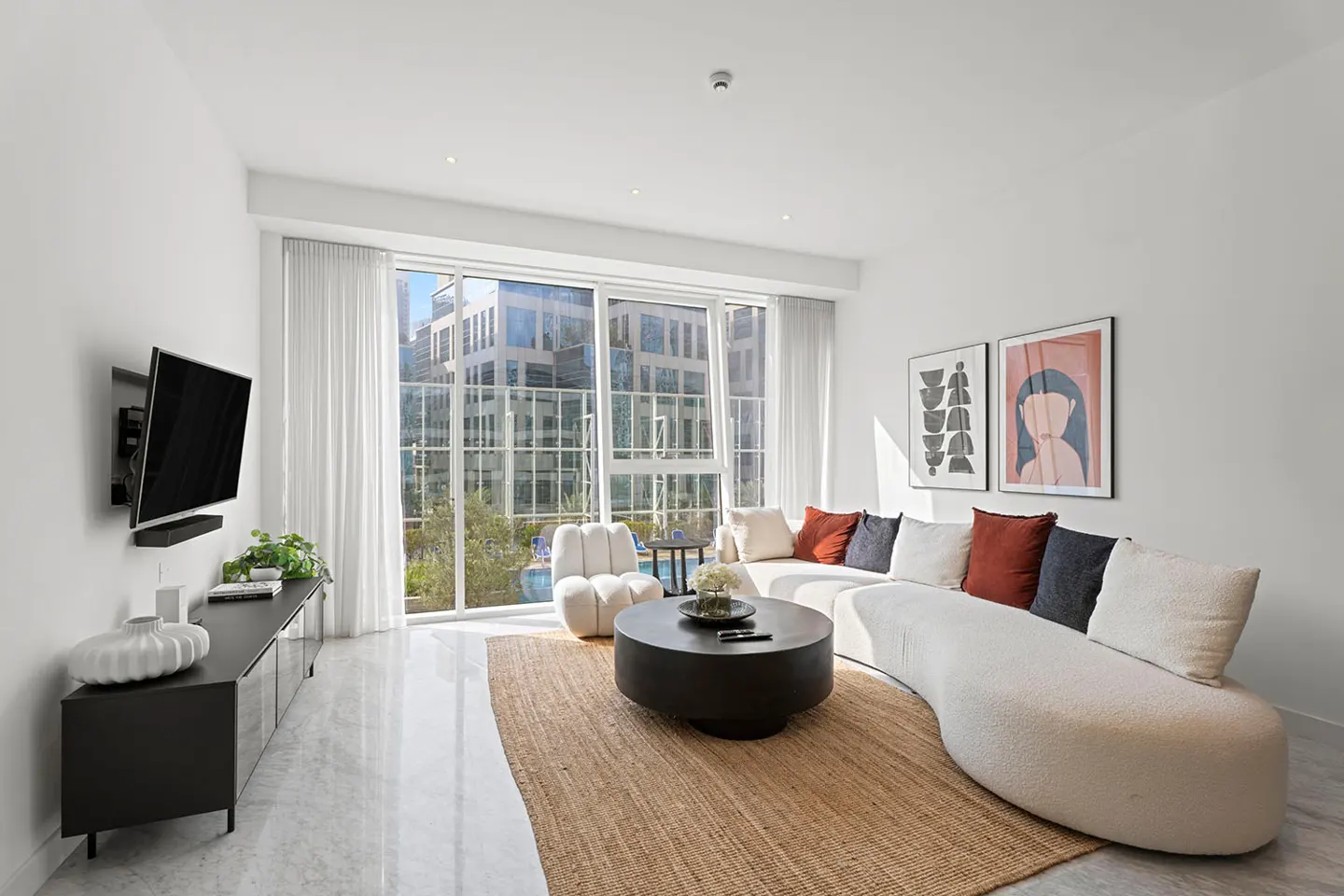 Bright living room with white walls, a curved white sofa with colorful pillows, and a round black coffee table on a jute rug. A TV is mounted on the wall.