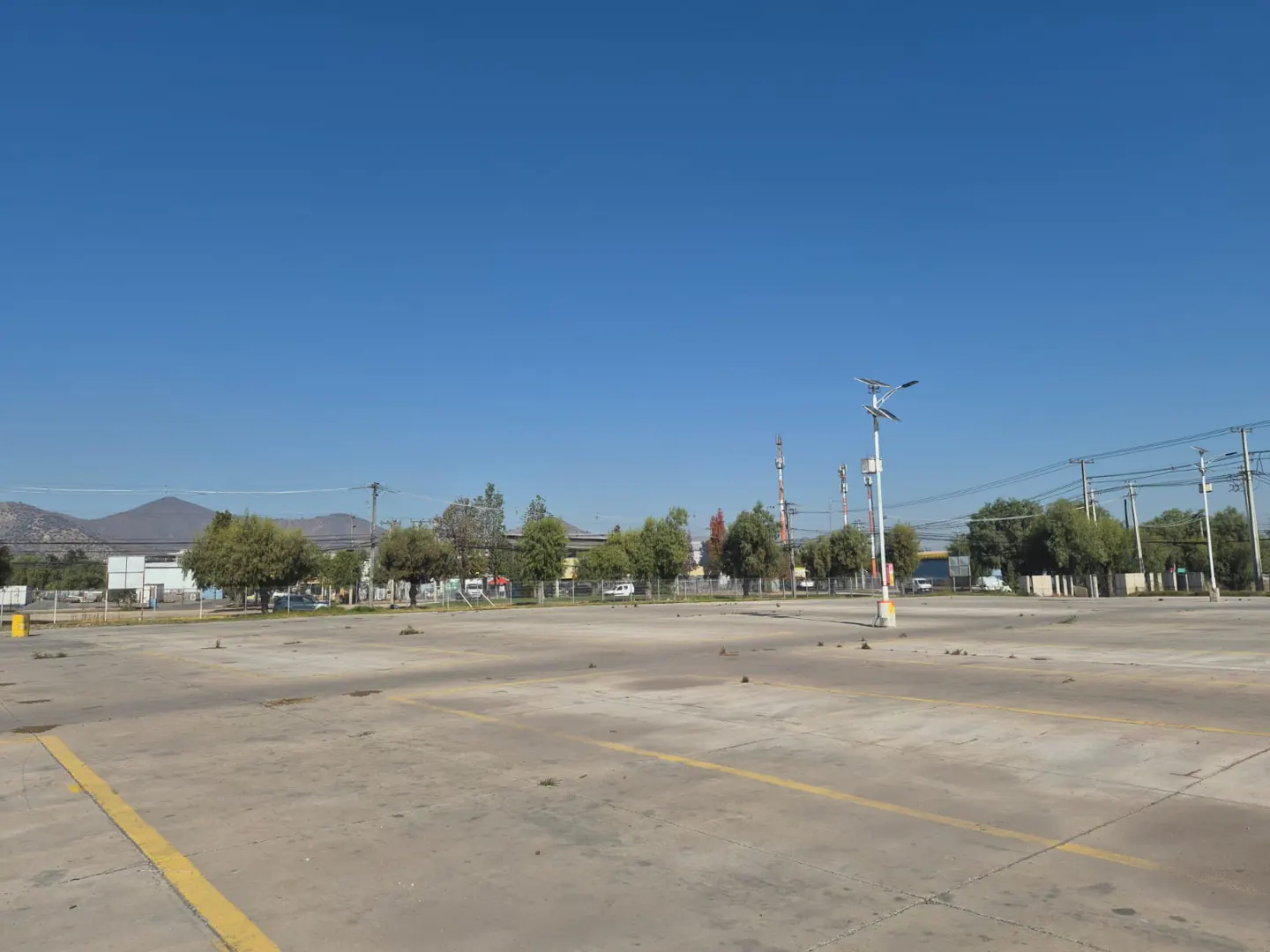 A wide, empty concrete parking lot under a clear blue sky, with distant trees, utility poles, and mountains in the back.