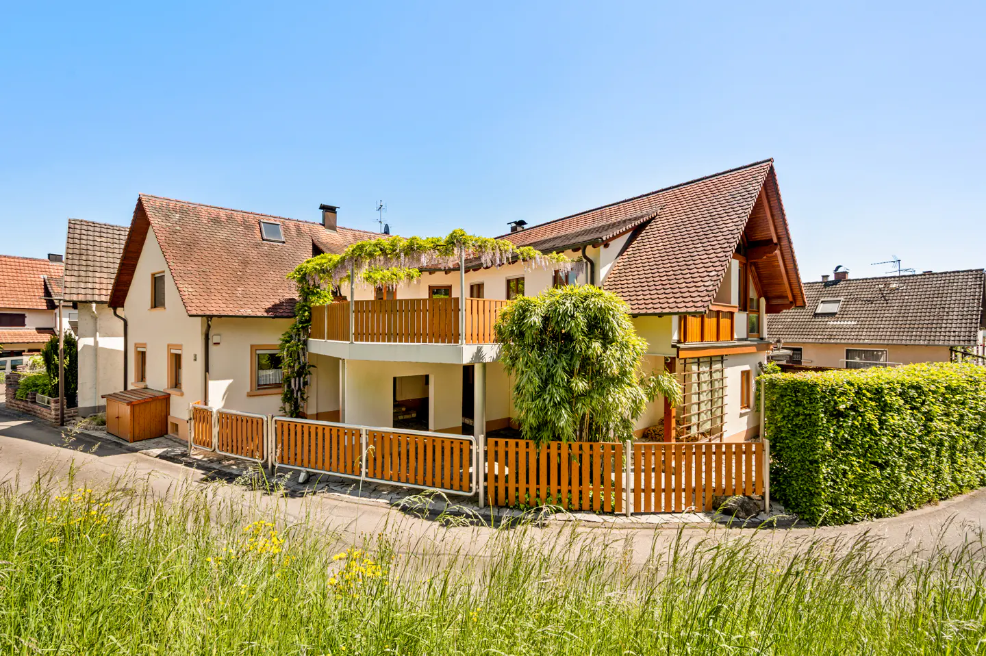 Two-story house with a brown roof and a wooden fence, surrounded by green grass and bushes. A balcony with wisteria is on the second floor.