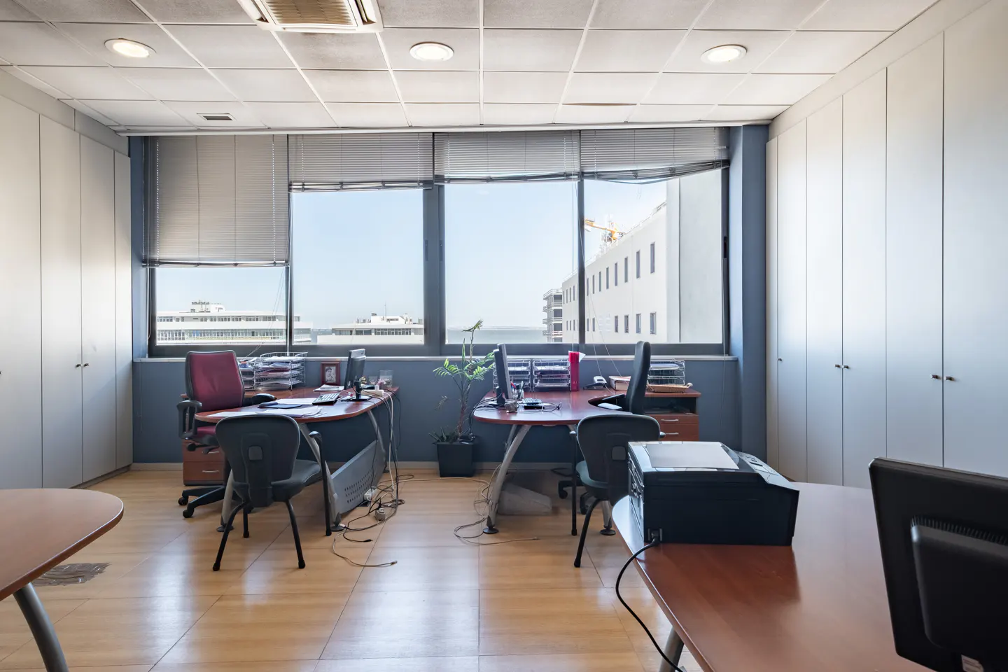 Bright office with wood floors, two desks, black chairs, built-in white cabinets, and large windows with city views.