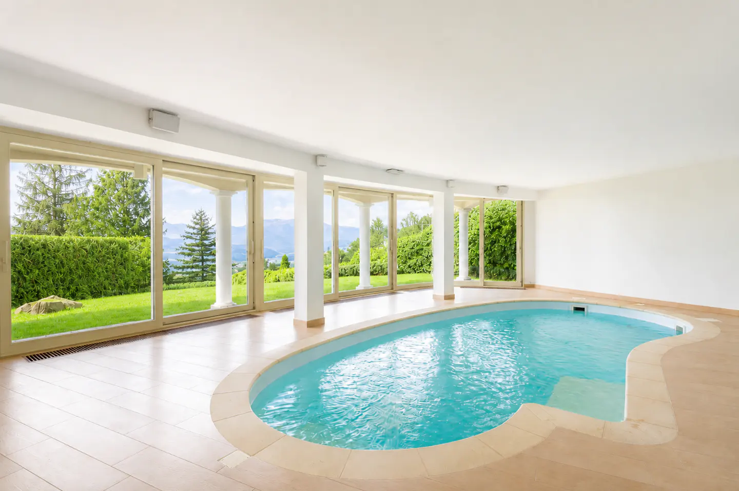 Indoor kidney-shaped swimming pool with light tile flooring, surrounded by large glass doors overlooking a green garden.