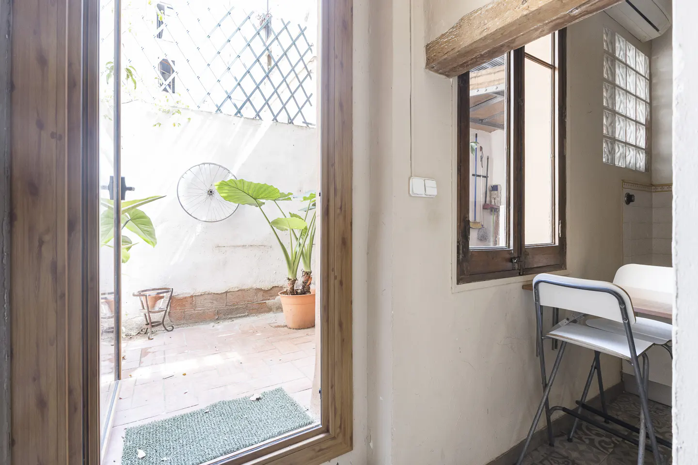 Interior view of a room with an open door leading to a small patio. The patio has plants, a white wall, and a bicycle wheel decoration. A window and table with chairs are visible inside.