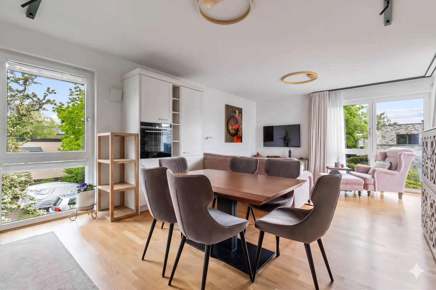 Bright, modern apartment interior with a wood dining table, grey chairs, and a pink armchair near a window.