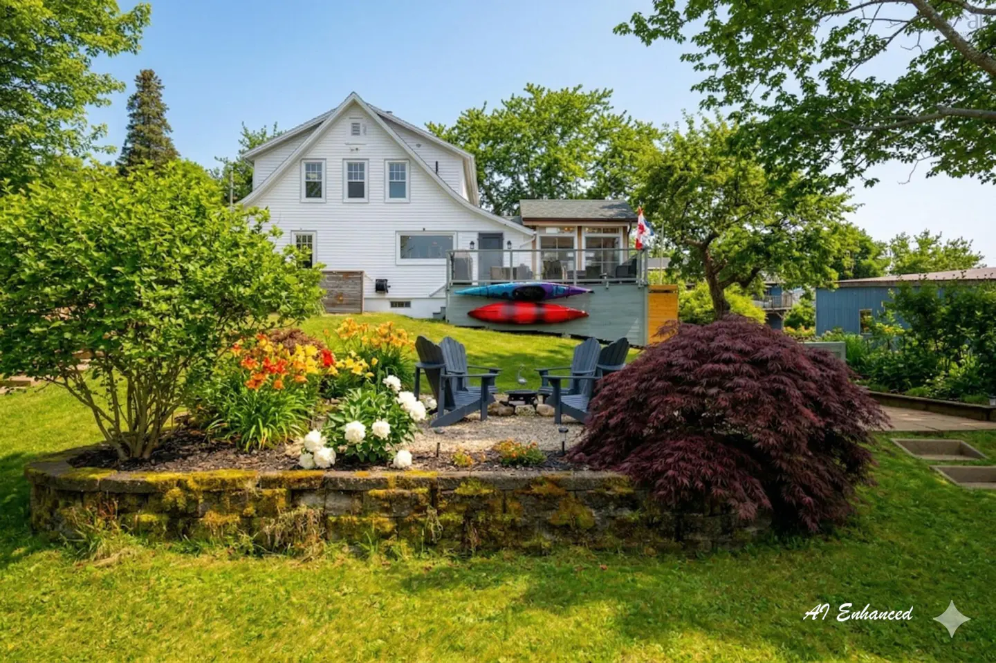 Backyard view of a white house with a deck, kayaks, and a fire pit with chairs. A flower bed adds color to the green lawn.