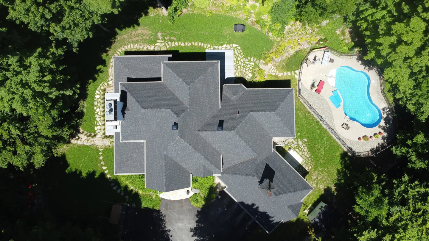 Aerial view of a large house with a dark gray roof, surrounded by green trees and grass, and a blue kidney-shaped pool.