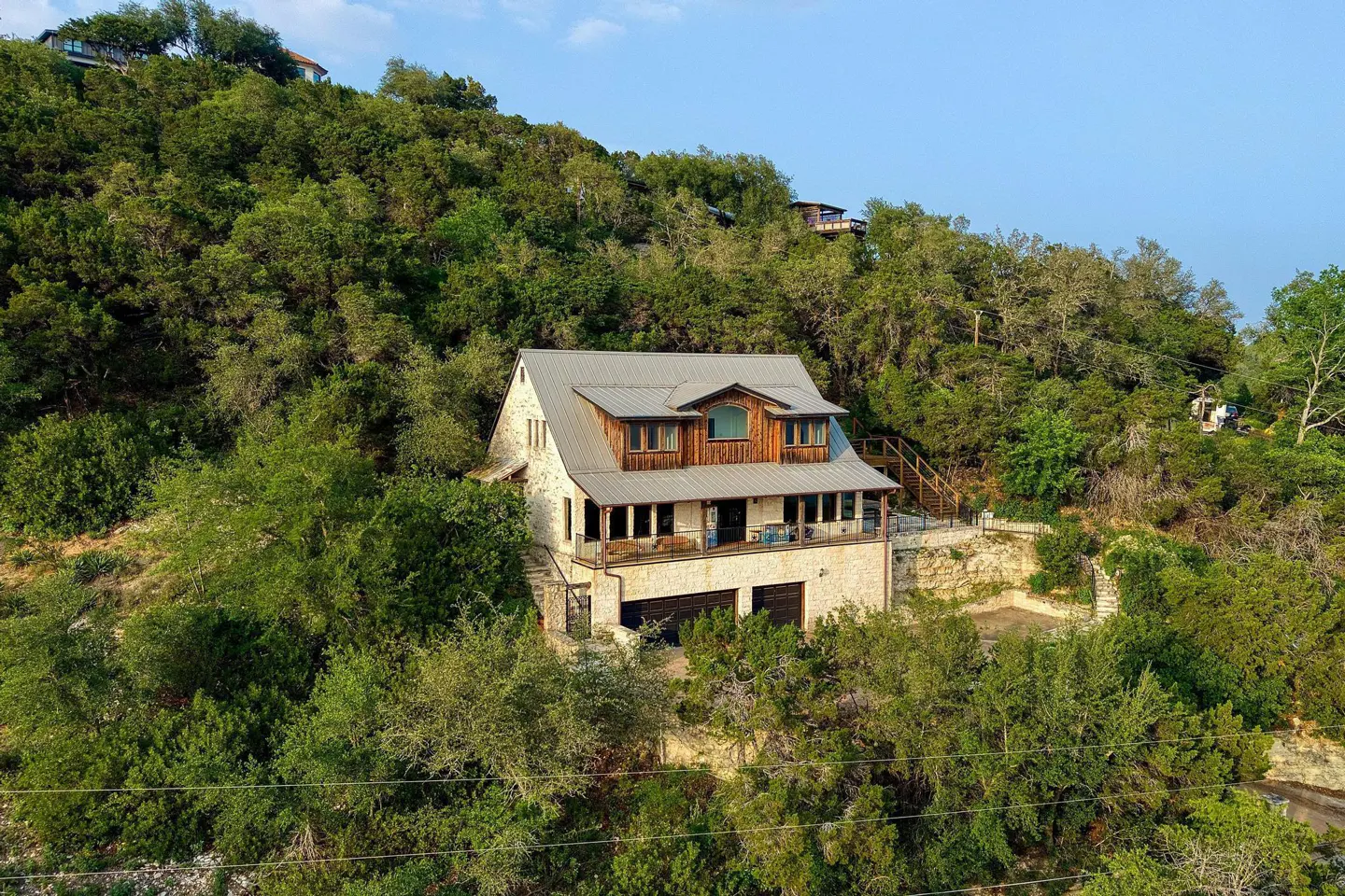 A stone house with a metal roof sits on a hill surrounded by green trees. The house has a two-car garage and a wooden deck.