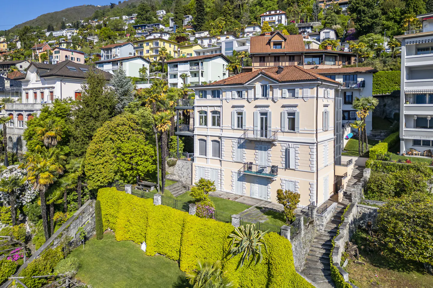 A three-story beige house with blue shutters sits on a green hillside with other houses in the background.