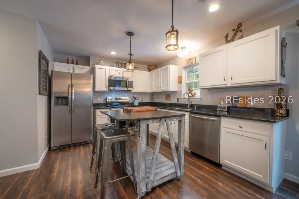 A kitchen with white cabinets, stainless steel appliances, and a dark wood floor. A rustic island with stools sits in the center.