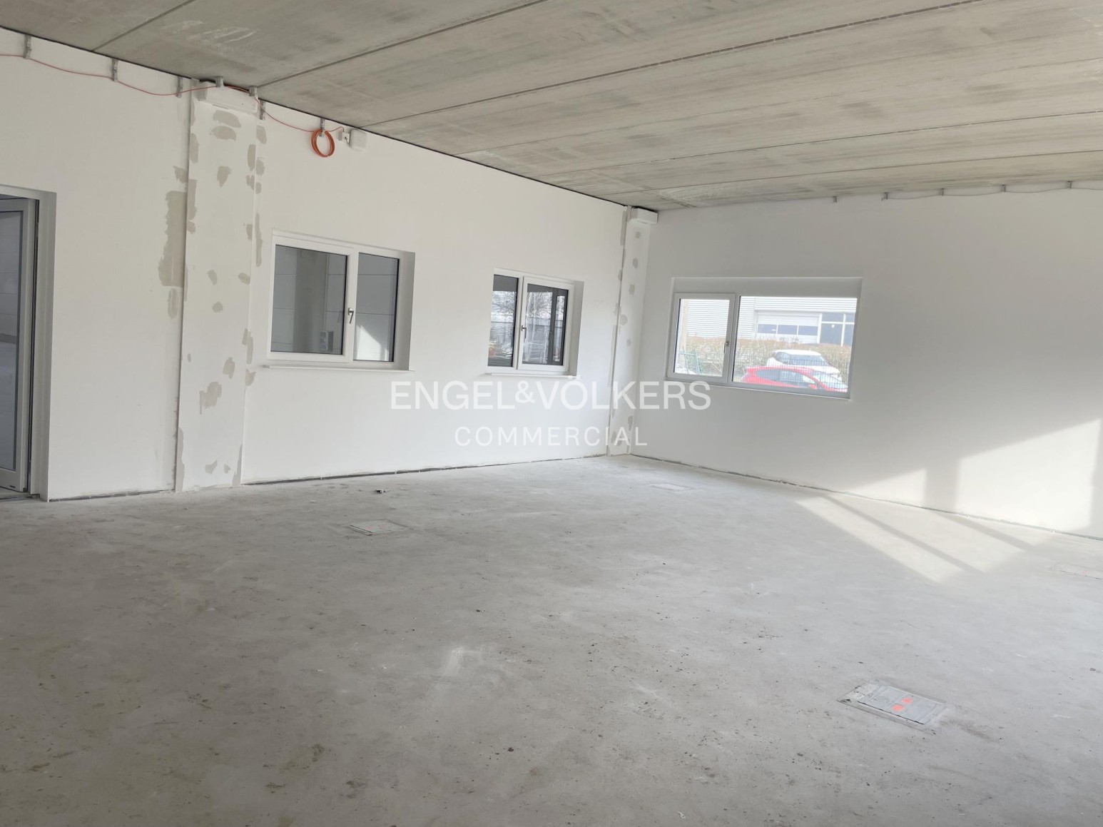 Unfinished commercial interior with white walls, concrete floor, exposed ceiling, and windows looking out to a parking lot. [AI-generated text]
