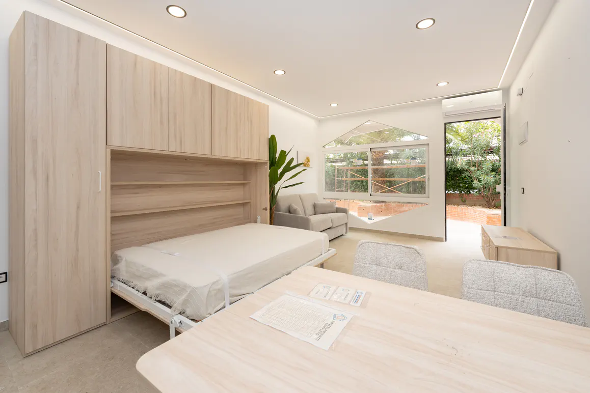 Bright studio apartment with a pull-down bed, light wood cabinets, a table with chairs, and a gray sofa. A large window and open door let in natural light.