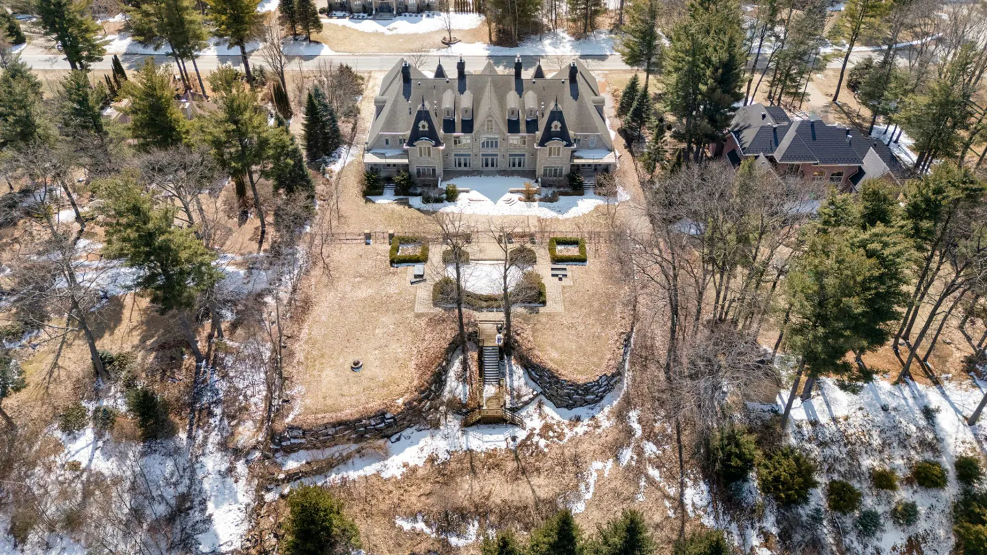 Aerial view of a large, grey stone mansion with black roofs, surrounded by trees and a lawn with snow patches.