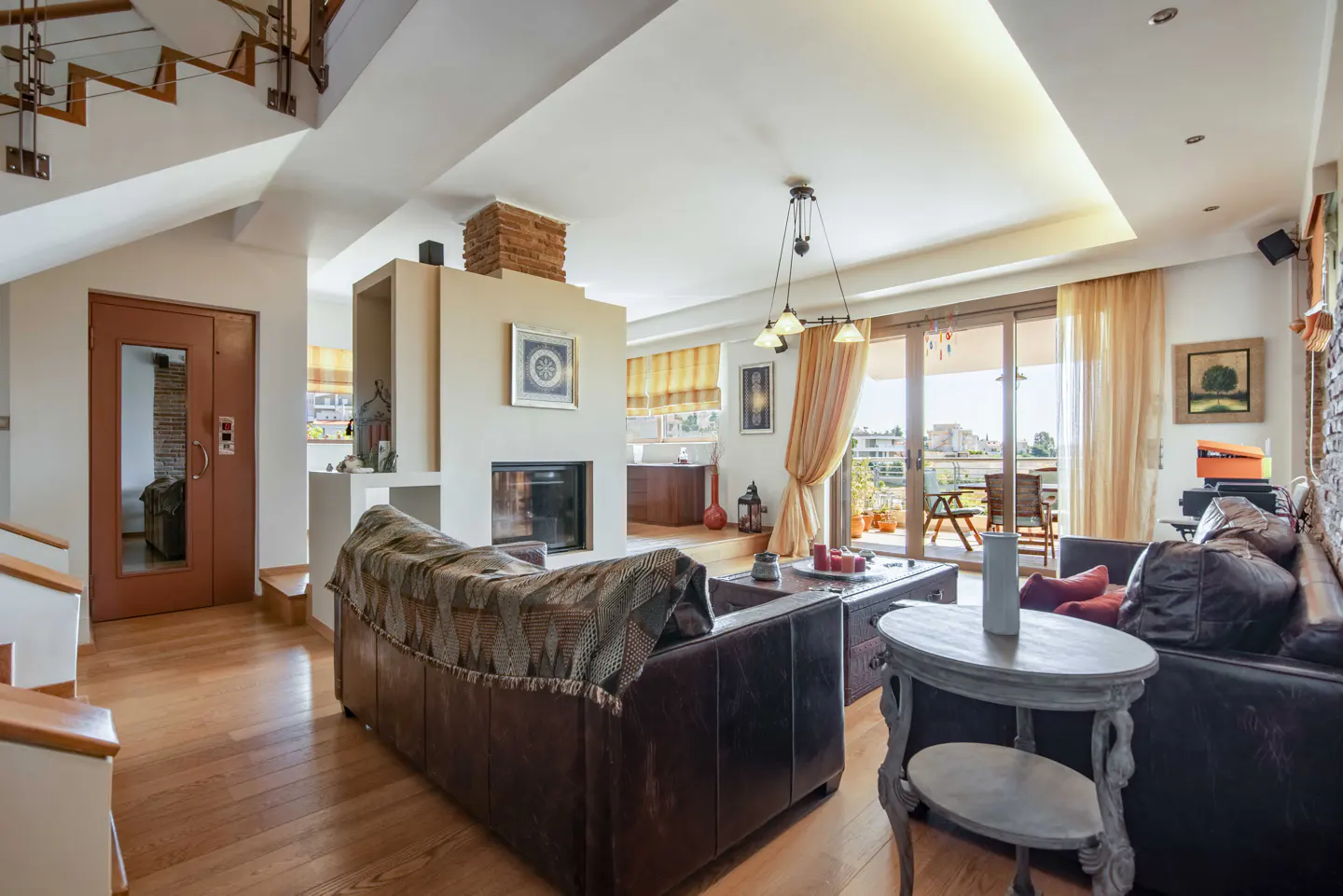 A bright living room with hardwood floors, a brown leather sofa, and a fireplace. A balcony is visible through sliding glass doors.
