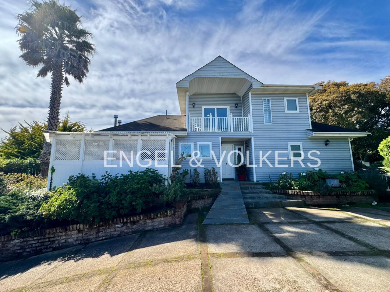 Two-story light blue house with a white balcony, lattice fence, and concrete driveway under a bright blue sky. [AI-generated text]