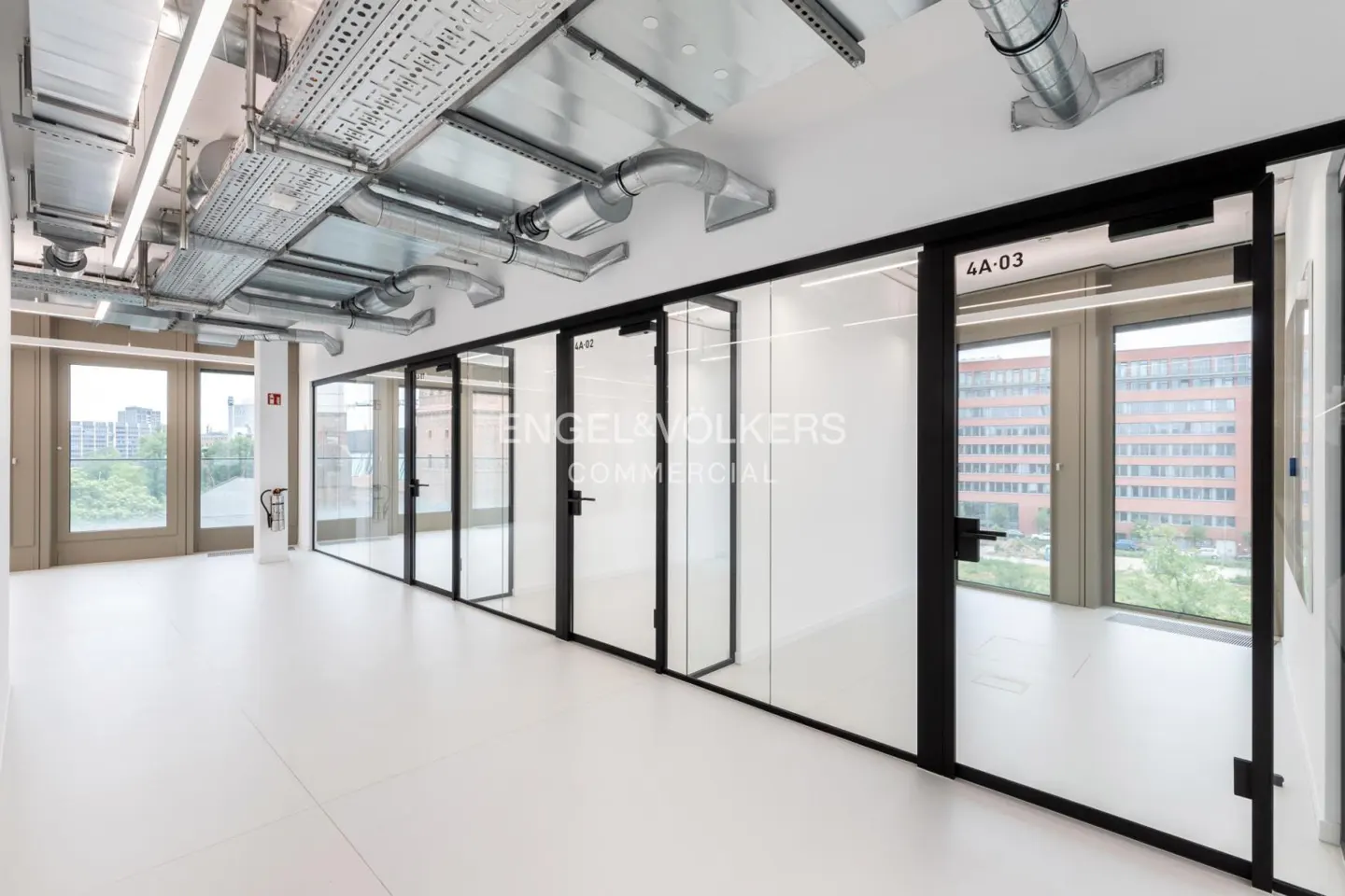 Bright office space with white floors, exposed ceiling pipes, and glass-walled offices with black frames. Windows overlook a cityscape.